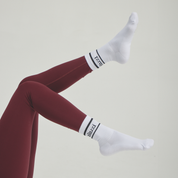 Woman wearing white Aluzi socks with black stripe details and logo, paired with leggings in a clean minimalist setting.
