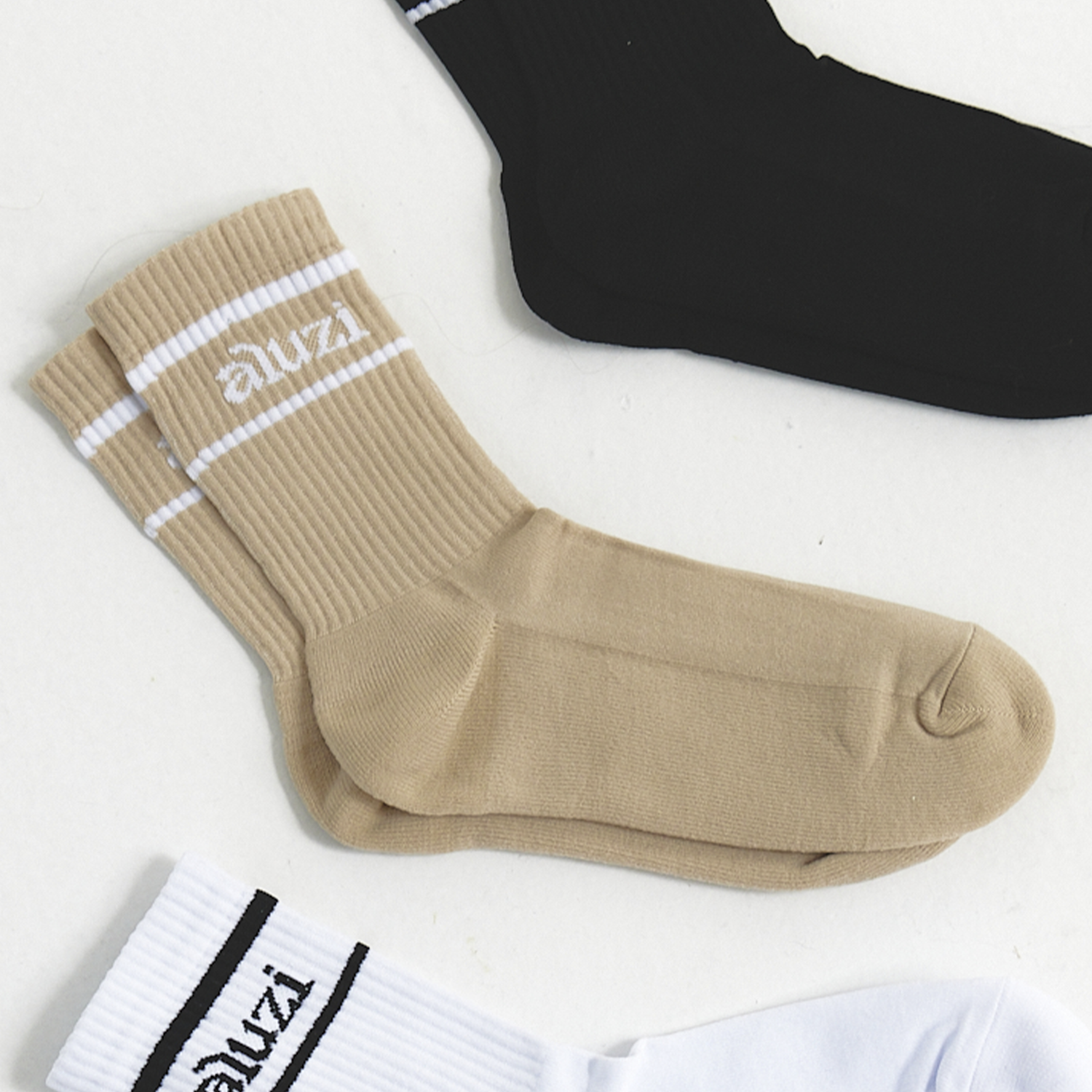 Beige ribbed crew socks with white stripes and “aluzi” logo, displayed on a white surface alongside black and white socks.
